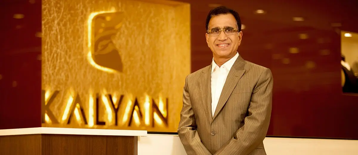 A suited man standing beside a KALYAN sign and logo in an elegant showroom setting.