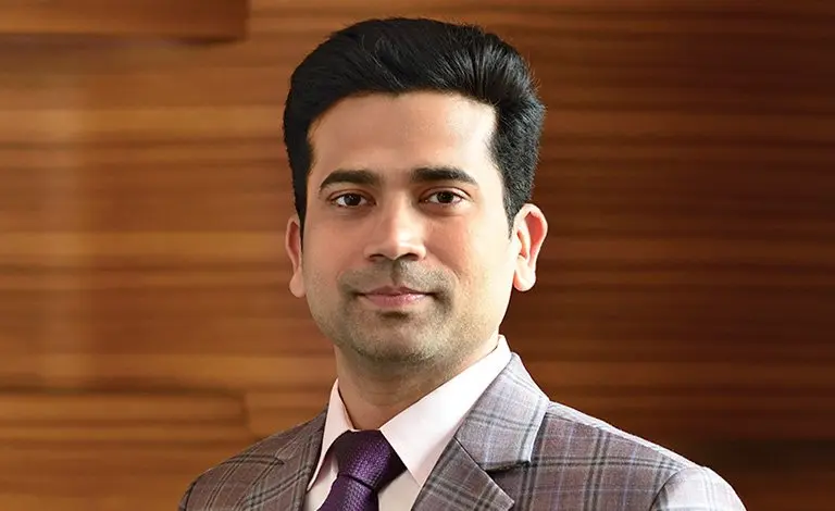 Professional portrait of a man wearing a gray checked suit, white shirt, and purple tie, standing against a wooden background.